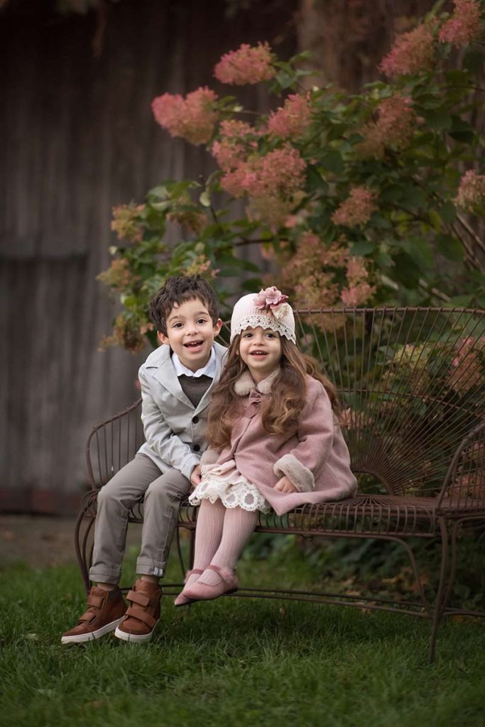 Two beautifully dressed children smiling on a wrought iron bench in a flower garden in Armonk, NY.