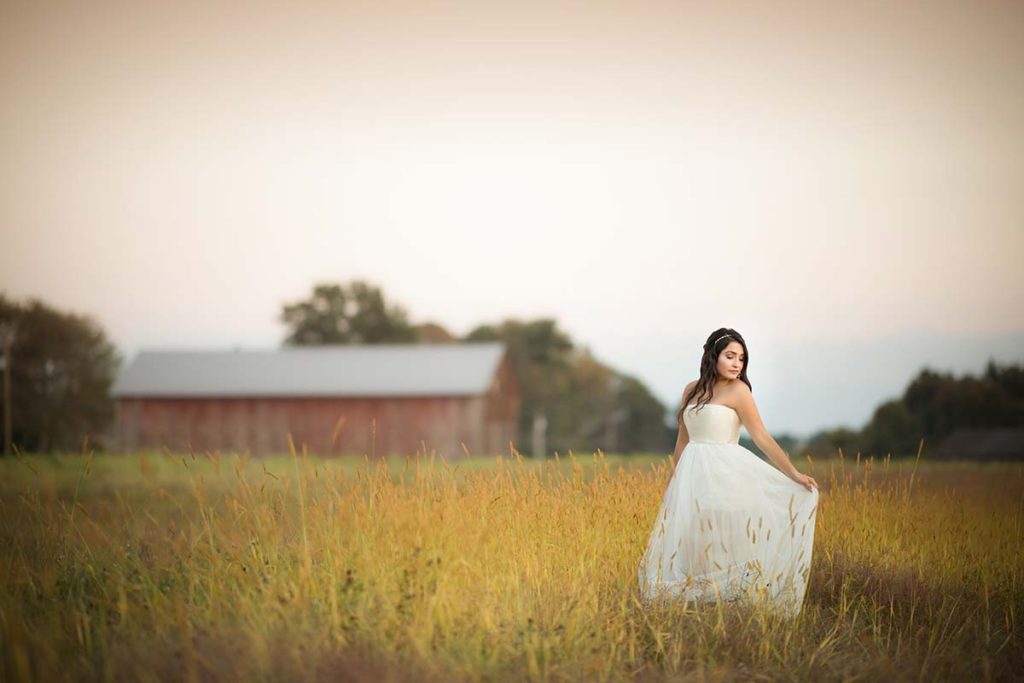A girl in a long white dress standing in a field of grass near a farm in Westchester county, NY.