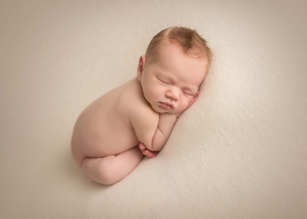 Infant baby with red hair sleeping on a beige blanket in this newborn photo