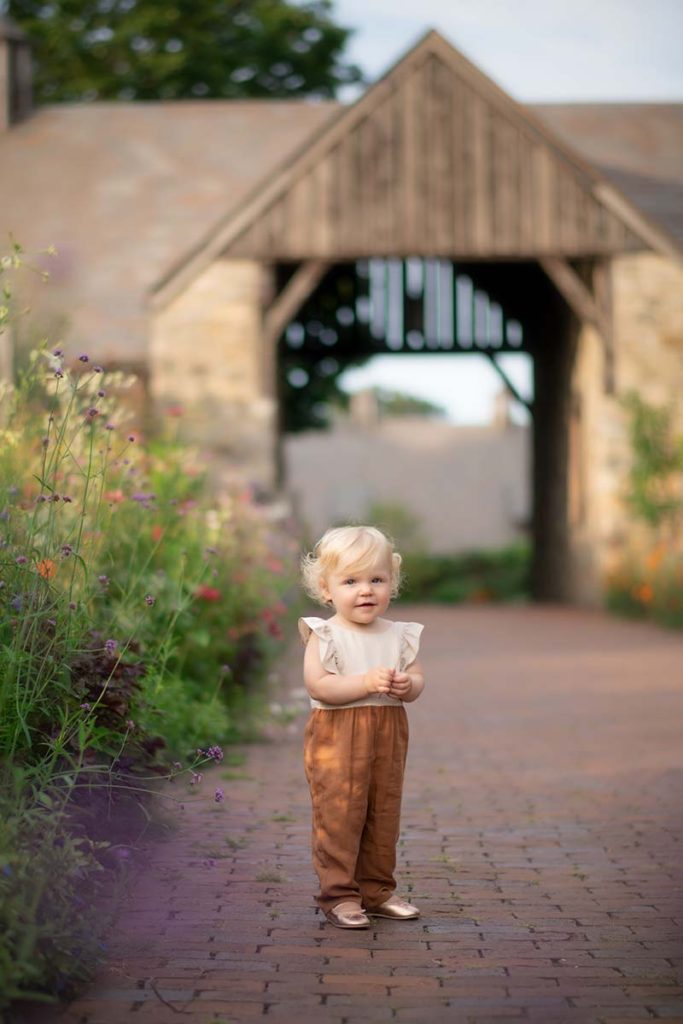 Happy baby with blonde hair standing on a brick path at a farm near Rye, NY.