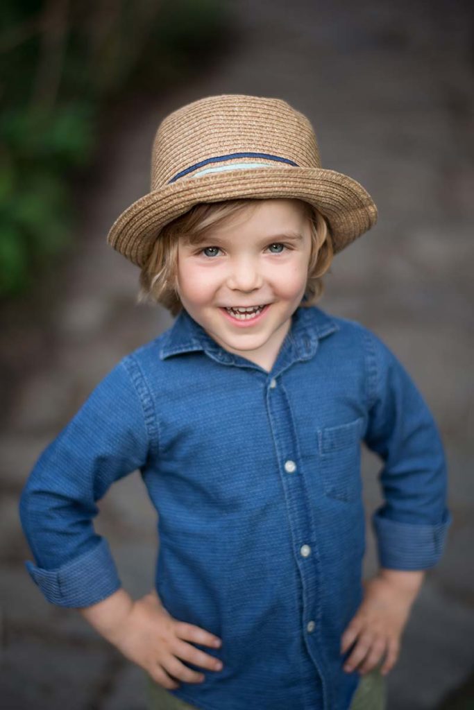 Handsome boy with blonde hair, blue shirt and a fedora hat smiling happily at the baby photographer.