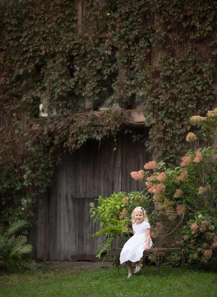 Young girl with blonde hair wearing a white dress sitting on a wrought iron bench in a flower garden on a Connecticut farm.