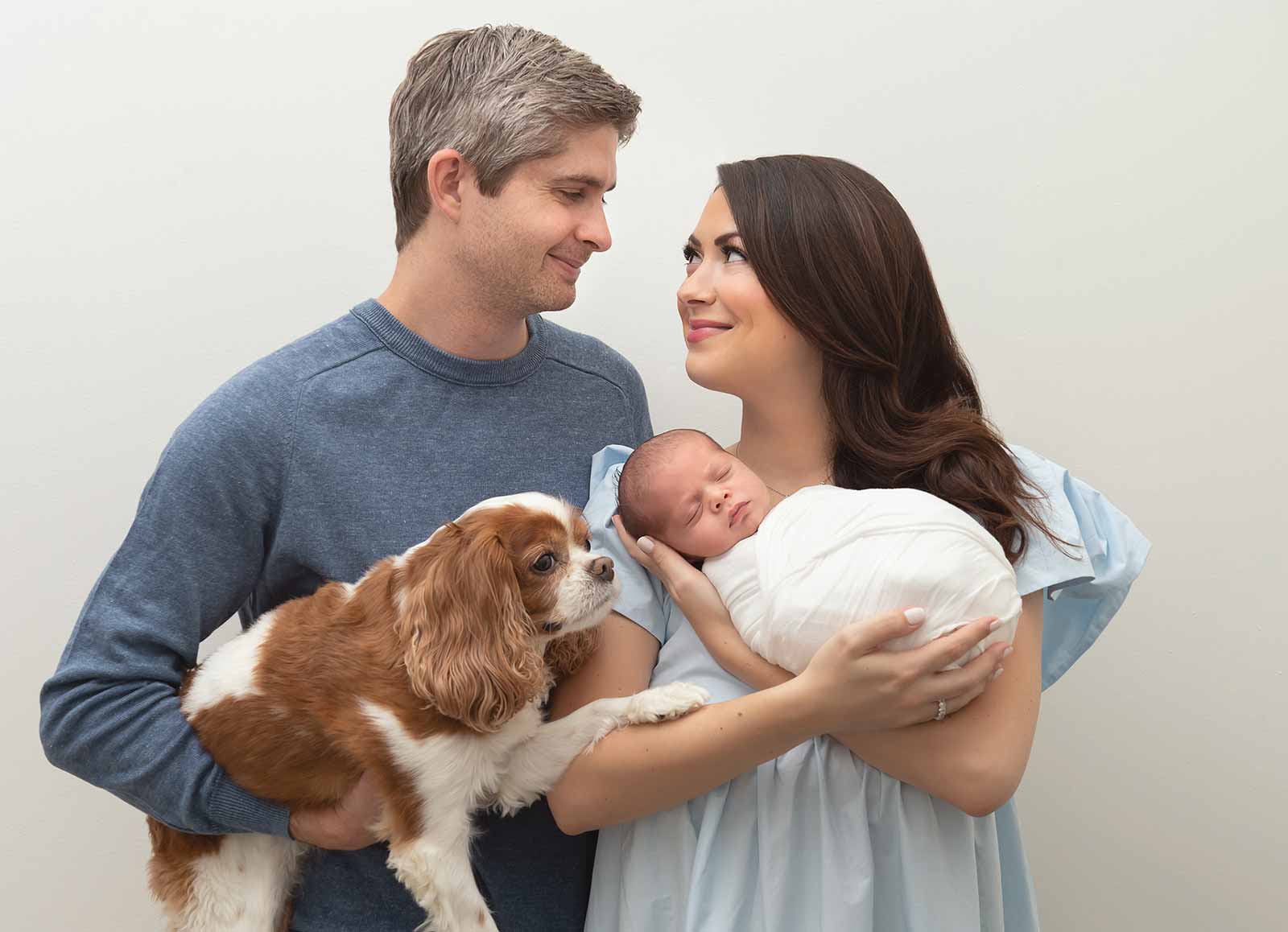 Mother and father looking at each other while holding dog and newborn