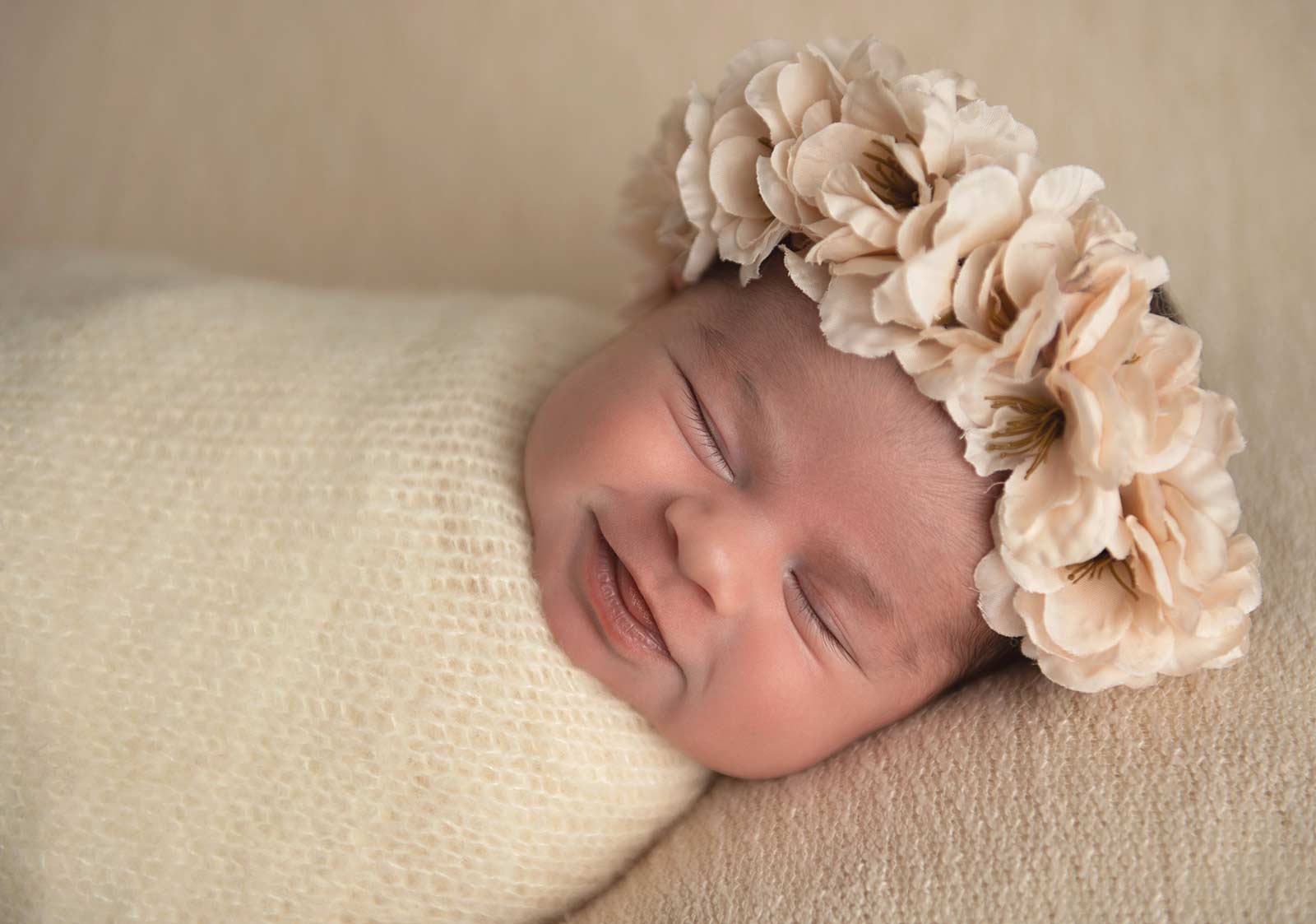 Sleeping newborn in beige knits and flower headband smiling