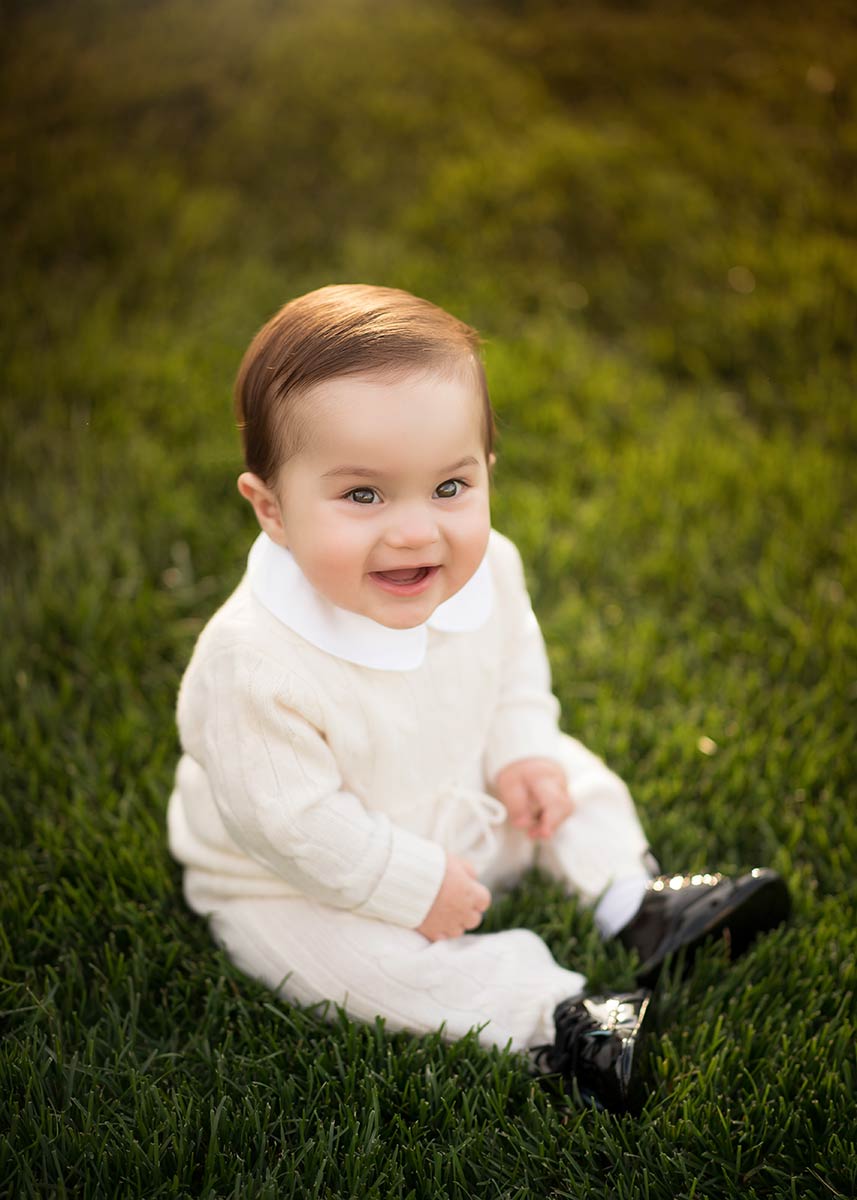 Adorable baby boy sitting down in grass and looking up at photographer