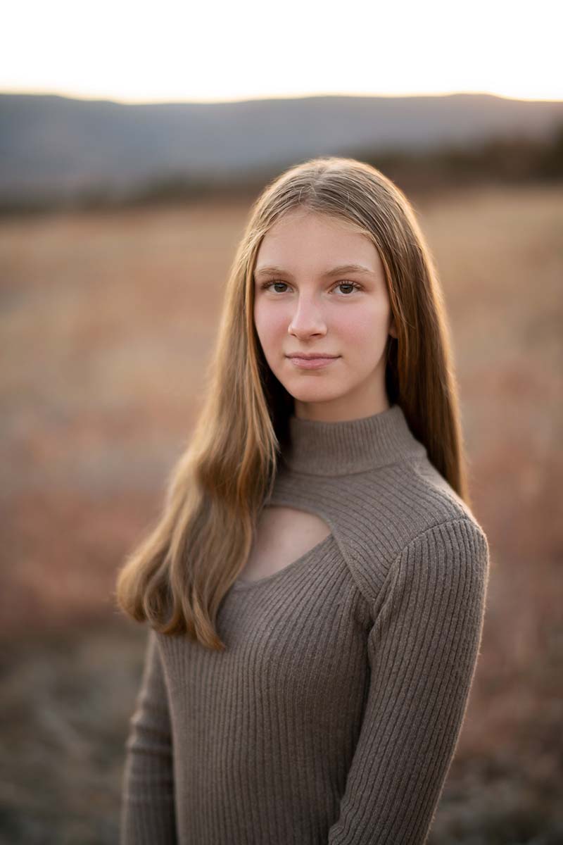 A young lady with long, straight hair stands outdoors with a neutral expression. She is wearing a ribbed, long-sleeved, taupe top with a keyhole neckline. The background is a blurred landscape, likely a field or plain, with soft, warm lighting.