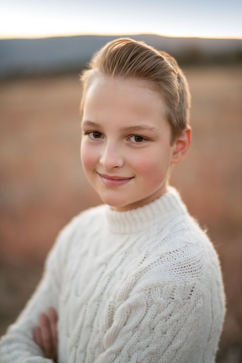 A young man with short, light hair smiles slightly at the camera. They are wearing a white, cable-knit sweater and standing outdoors with an out-of-focus field and mountain range in the background, softly lit by natural light. Captured through the lens of a talented Denver photographer.